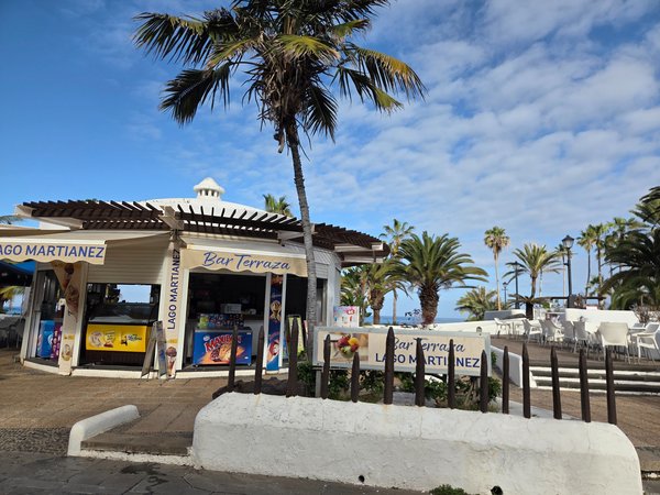 Bar terraza del Lago Martiánez en Puerto de la Cruz, con palmeras, mesas al aire libre y vistas al océano Atlántico en un entorno turístico.