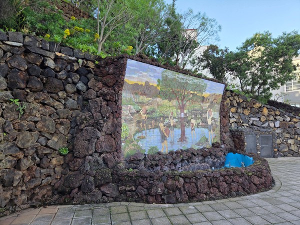 Mural cerámico incrustado en un muro de piedra volcánica en el Parque Taoro, con una pequeña pila azul y vegetación alrededor.