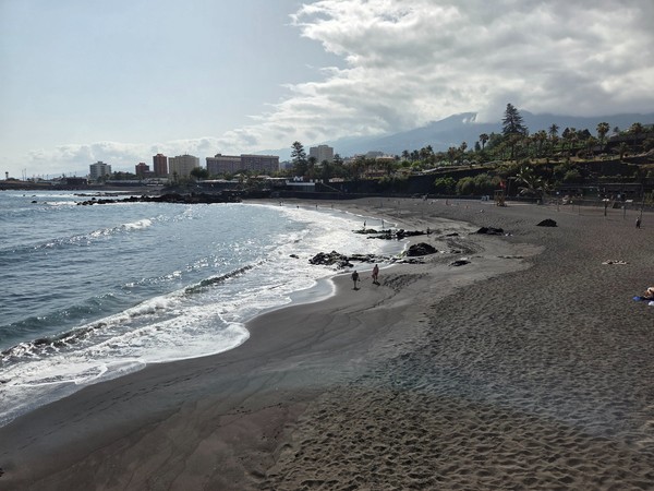 Vista panorámica de la playa de Punta Brava en Puerto de la Cruz, con arena volcánica negra, olas suaves y el Teide al fondo entre nubes.
