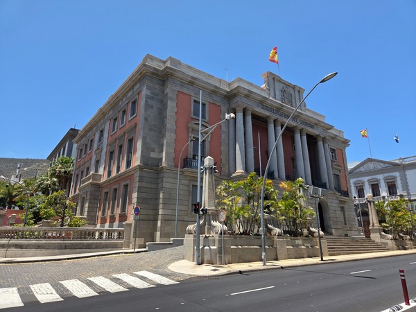 Edificio histórico de la Subdelegación del Gobierno en Santa Cruz de Tenerife, con columnas neoclásicas y banderas ondeando bajo un cielo azul.